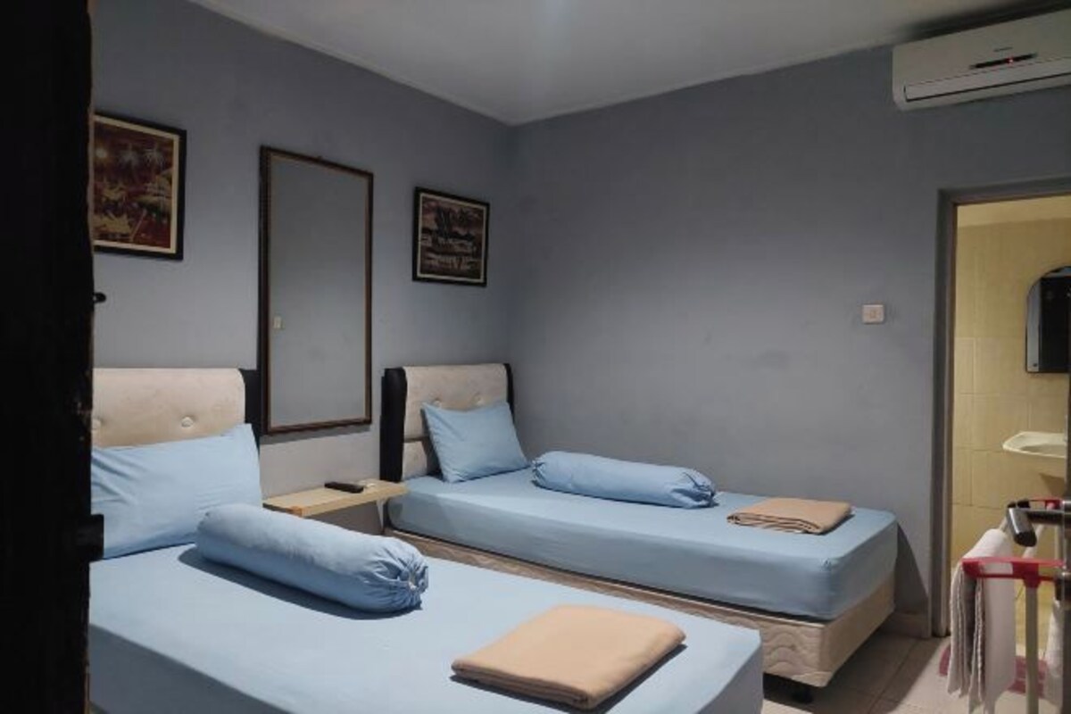 A room with two beds, each with a blue mattress and matching pillows. There is a wooden nightstand between the beds, and a beige blanket is folded on the bed in the foreground.