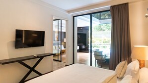 Una habitación de hotel moderna con una cama grande, un televisor de pantalla plana montado en la pared y un ventanal corredero que da a un balcón con mobiliario exterior.