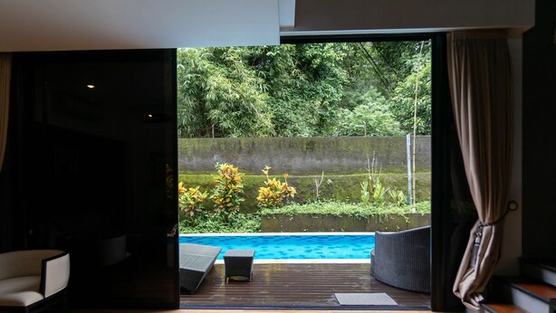 Una habitación moderna con suelo de madera, un amplio ventanal corredero que da a una piscina y vistas de abundante vegetación.