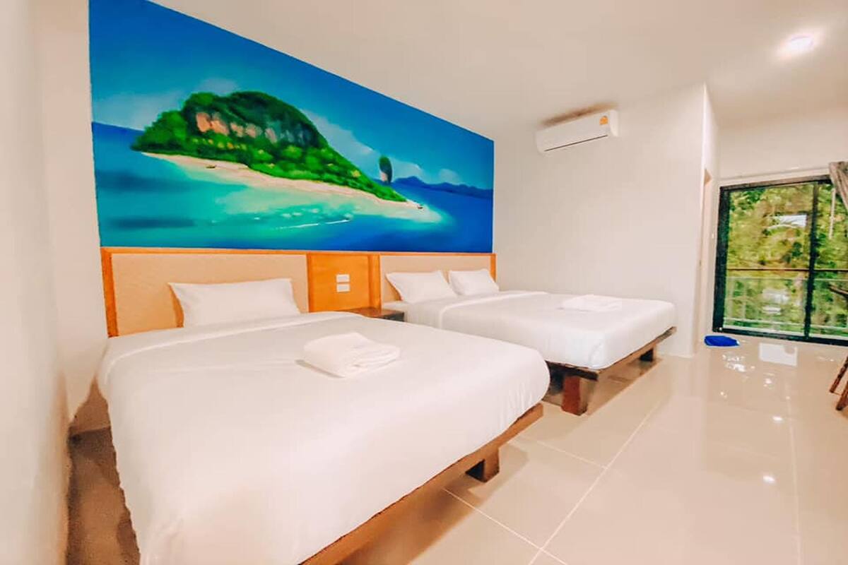 Quarto de hotel com duas camas, um quadro de uma ilha tropical e uma grande janela com vista para o verde.