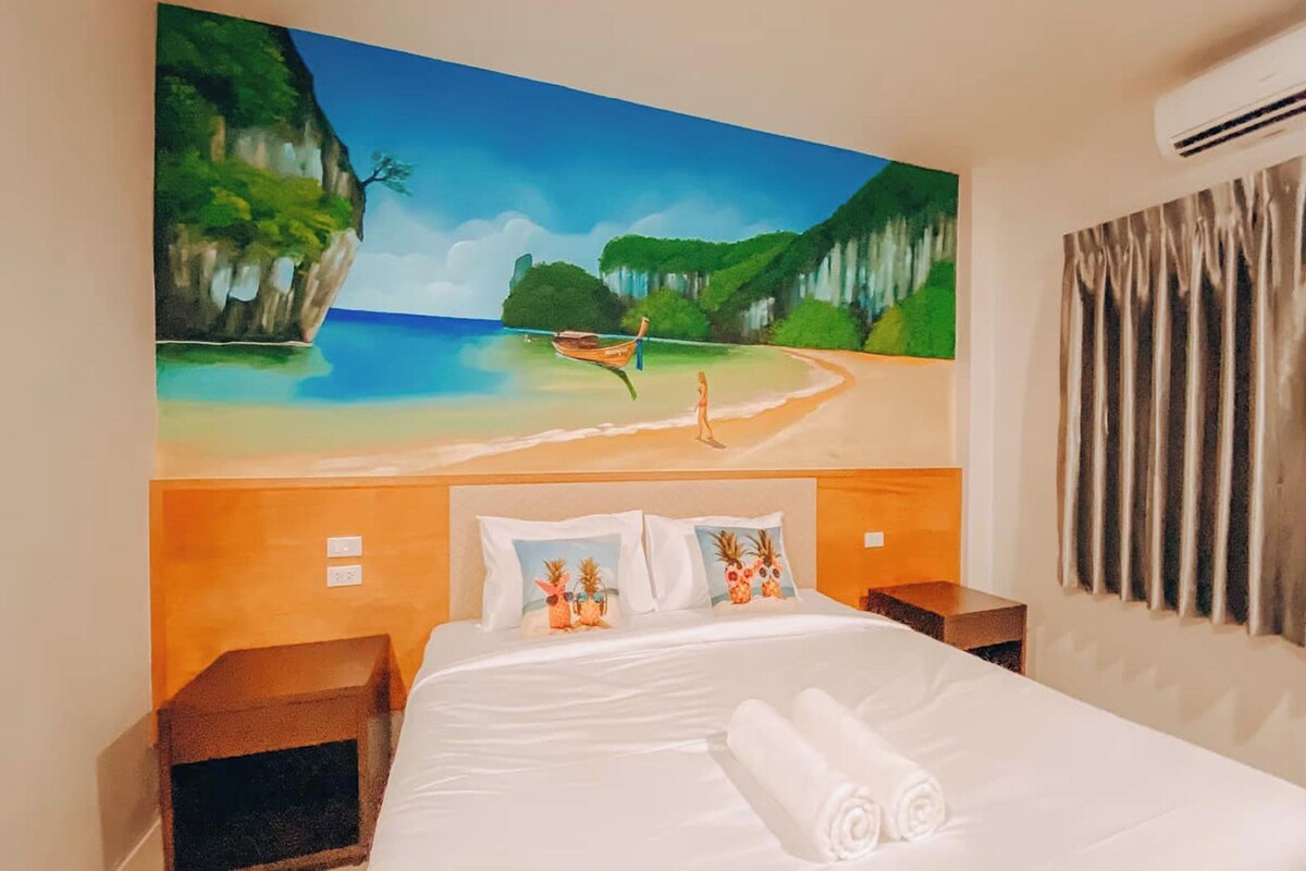 A hotel room with a large beach painting on the wall, a neatly made bed with white linens, and a window with curtains.