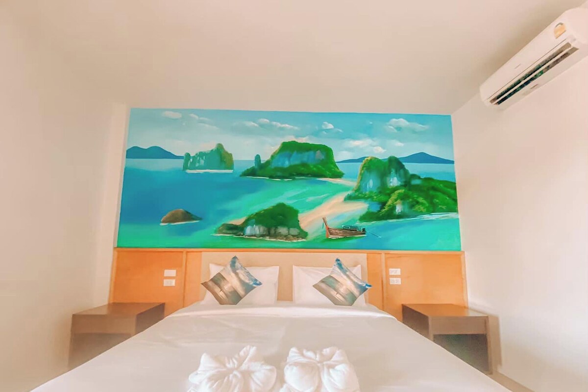 A hotel room with a bed, a painting of a tropical landscape, and a small boat on the water.