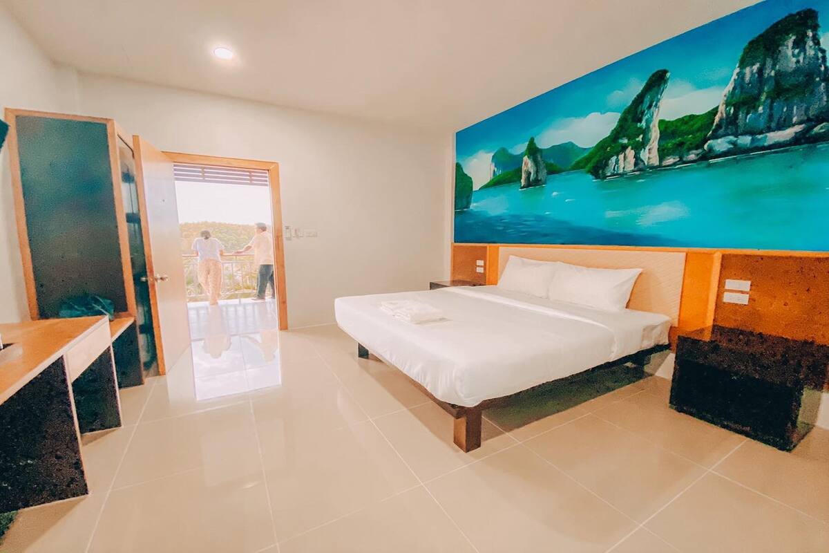 Quarto de hotel com cama, escrivaninha e pintura de uma paisagem costeira.