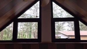 Una habitación con dos ventanas grandes, cada una con un marco triangular, que ofrece vistas a un área boscosa y un edificio de techo rojo.
