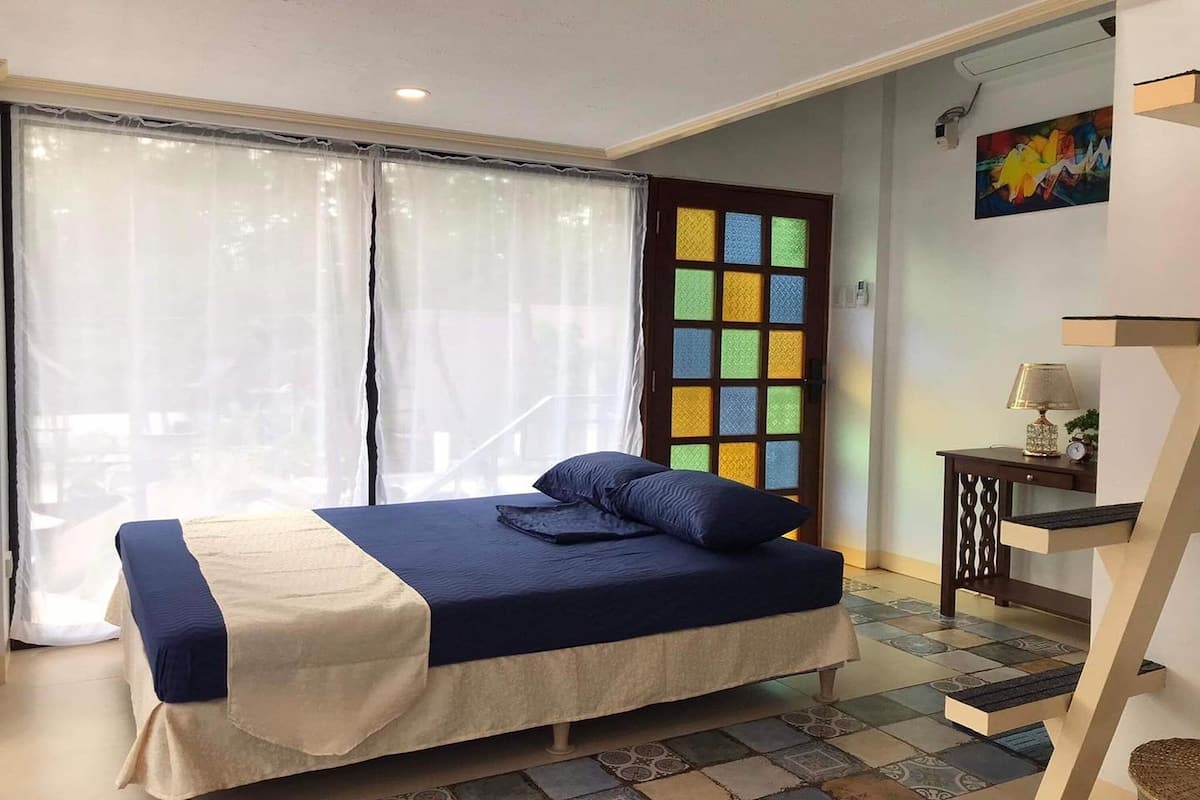 Une chambre à coucher avec un lit, une fenêtre avec des rideaux, une porte avec des panneaux de verre colorés, une petite table avec une lampe et un escalier.