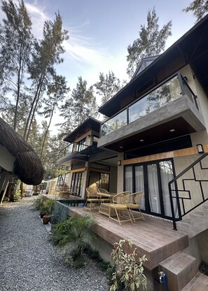 A modern two-story house with a patio area, outdoor seating, and a view of tall trees.