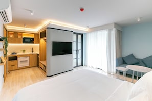 A modern hotel room with a large bed, a TV, a kitchenette, and a seating area with a sofa and two small tables.
