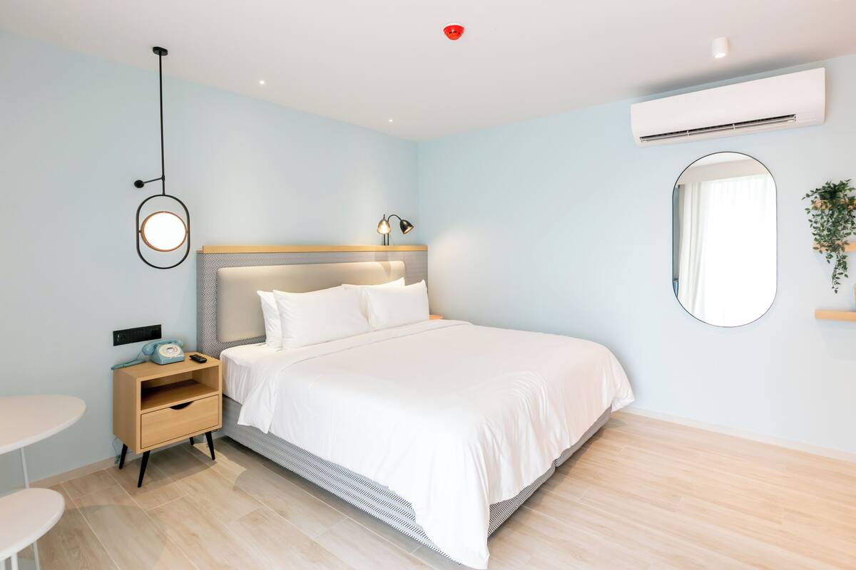 A modern hotel room with a bed, bedside table, lamp, and a round mirror on the wall.
