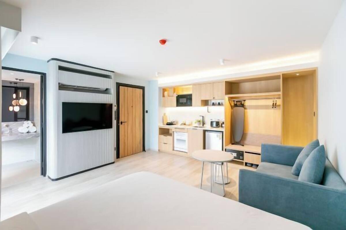 A modern room with a bed, a small table, a sofa, a TV, and a kitchenette.