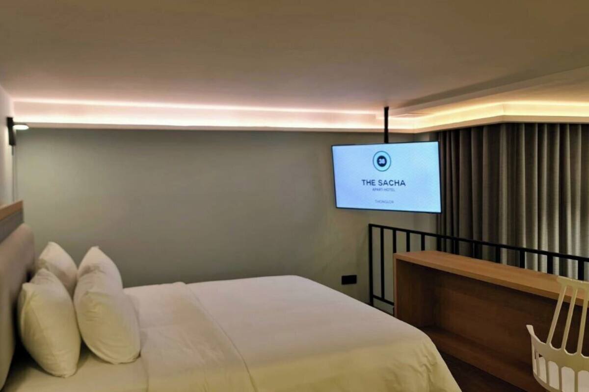 A hotel room with a bed, a TV mounted on the wall, a wooden headboard, and a window with curtains.