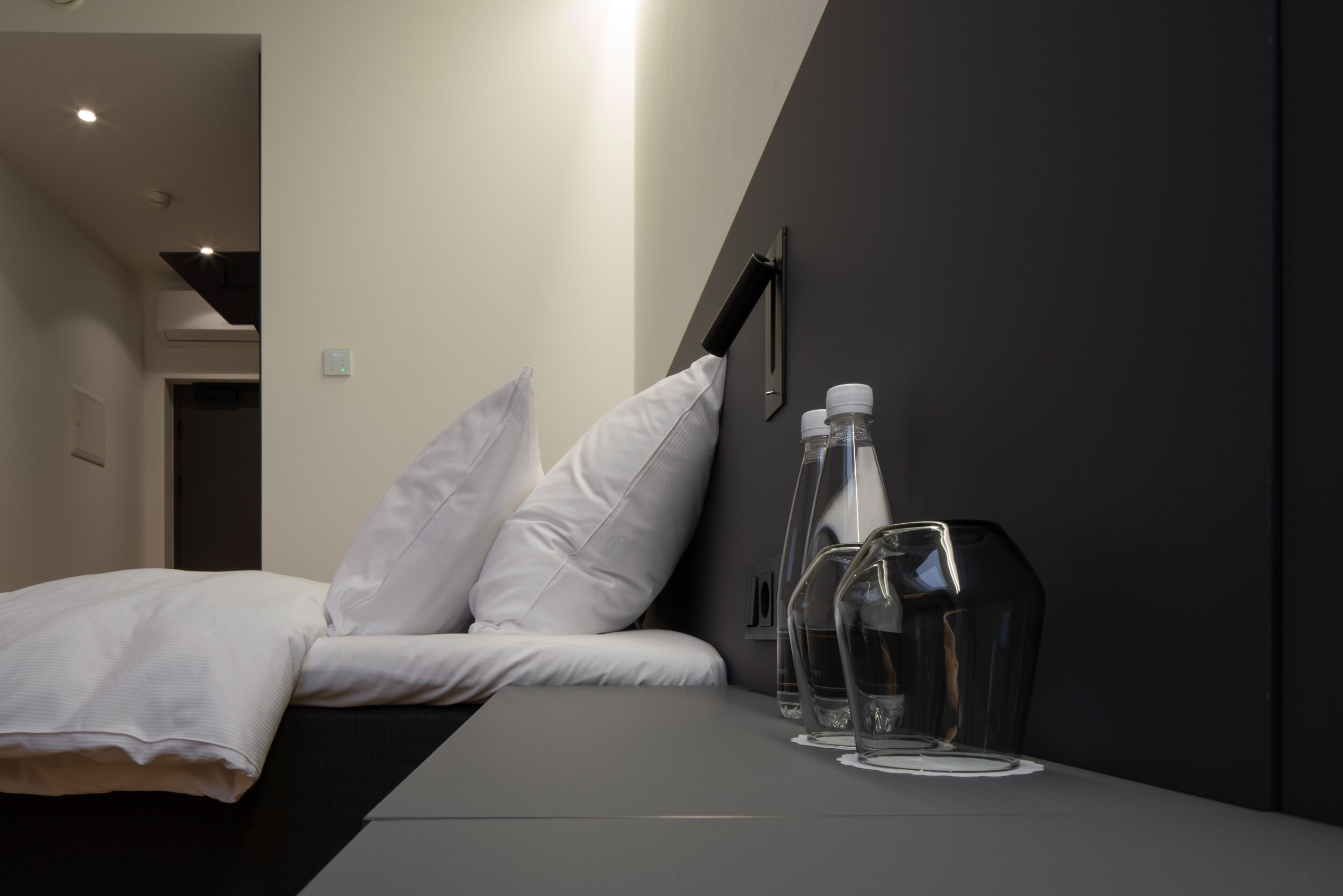 Camera d'albergo moderna con un letto, cuscini bianchi, un comodino grigio e due bottiglie di vetro trasparente.