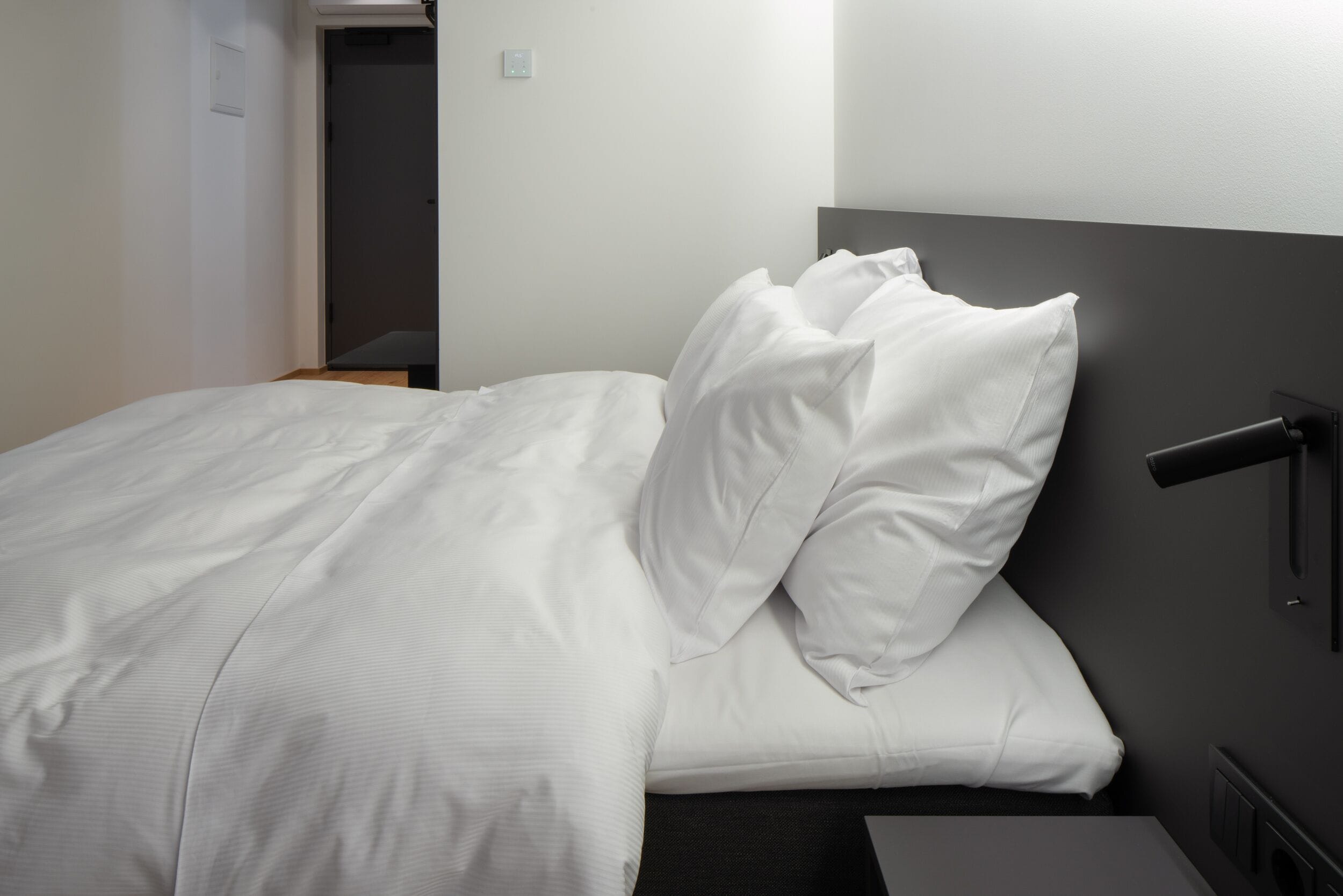 Una cama bien tendida con sábanas blancas, cabecera oscura y mesita de noche con lámpara.