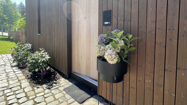 A wooden building with a stone-paved entrance, a hanging planter with flowers, and a doorbell button. - New vacation cottage in the idyllic Ilz valley. 5 minutes to Passau (Salzweg)