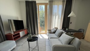 A modern living room with a sofa, a TV, a small table, and a balcony with curtains. - New vacation cottage in the idyllic Ilz valley. 5 minutes to Passau (Salzweg)