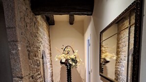 A hallway with stone walls, a tiled floor, a wooden table with a stack of books, a mirror, and a decorative vase with flowers. - Authentic 16th-century Breton house in the heart of downtown, 2 minutes from the beach (Saint-Pol-de-Léon)