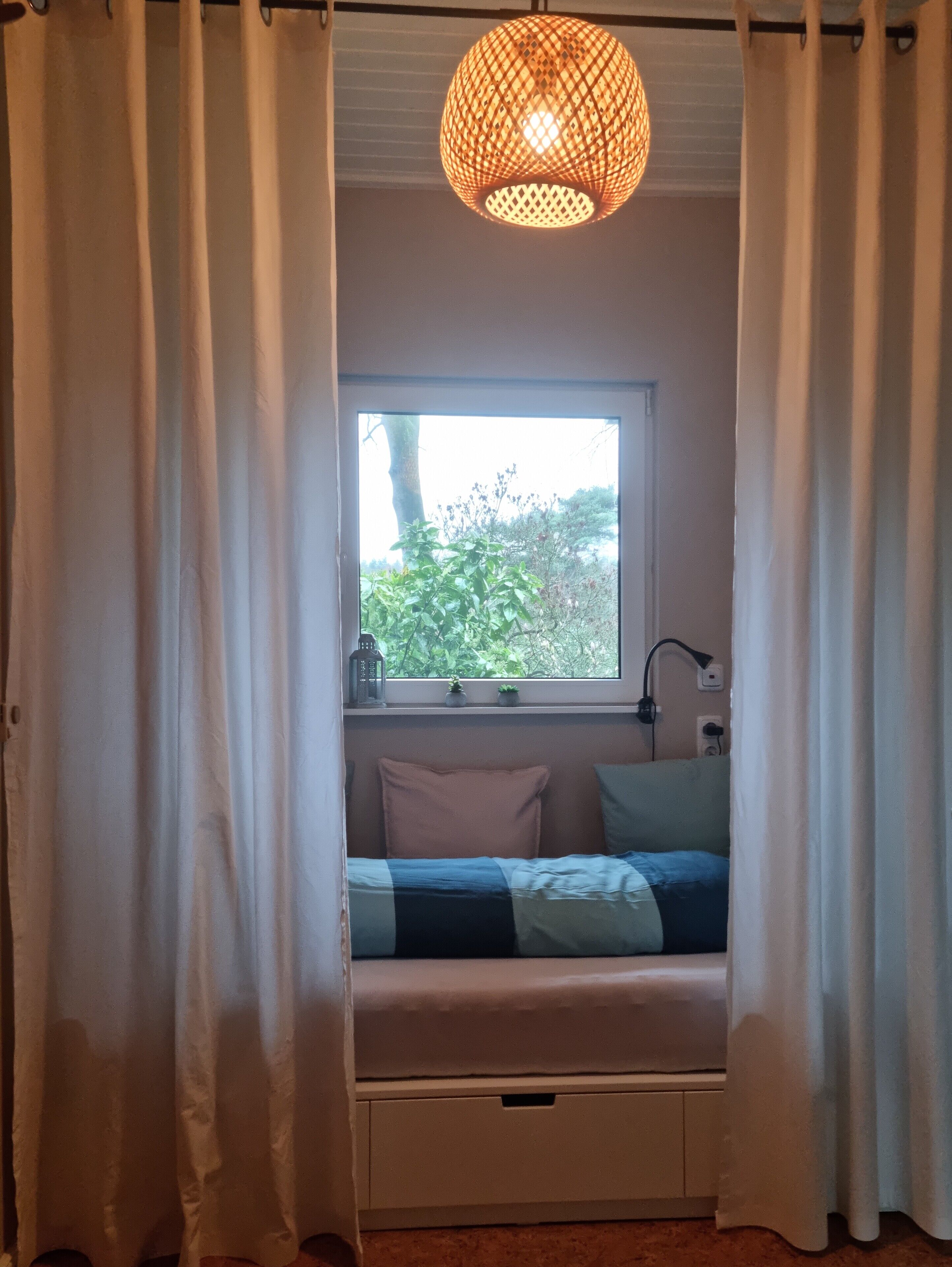 A bedroom with a window, a bed, a nightstand, a lamp, and a hanging light fixture.