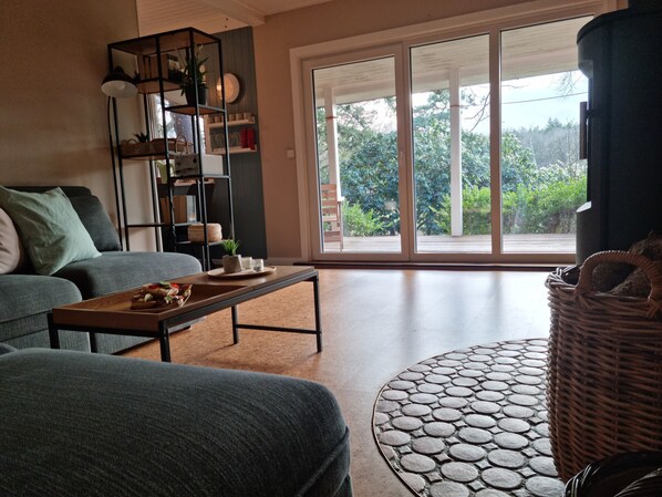 A living room with a grey sofa, a coffee table with a tray of food, a bookshelf, and a large sliding glass door leading to a balcony with greenery.