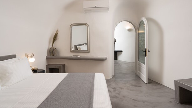 A modern bedroom with a bed, bedside table, mirror, and an arched doorway leading to another room. - Occhio Villas Santorini (Santorini)