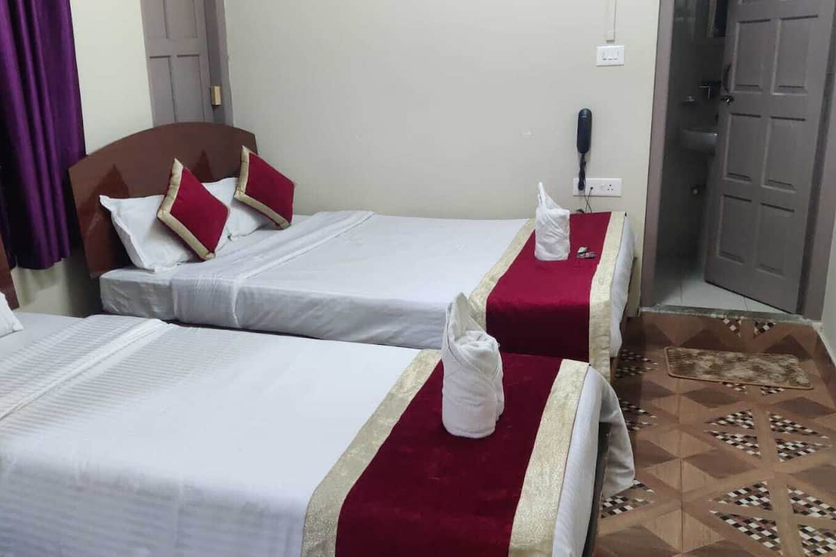 A hotel room with two single beds, each with white linens and red pillows. There is a red and gold bed runner on one bed and a white towel on the other. The room has a patterned floor and a door to the right.