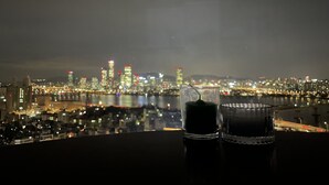 A night view of a city skyline with buildings and lights reflected in a body of water. Two glasses of a dark beverage are placed on a table in the foreground. - Amazing river view! best place to travel Seoul :) (서울특별시)