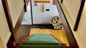 Un escalier en bois recouvert d’une moquette verte, une cheminée et un tableau représentant un paysage enneigé.