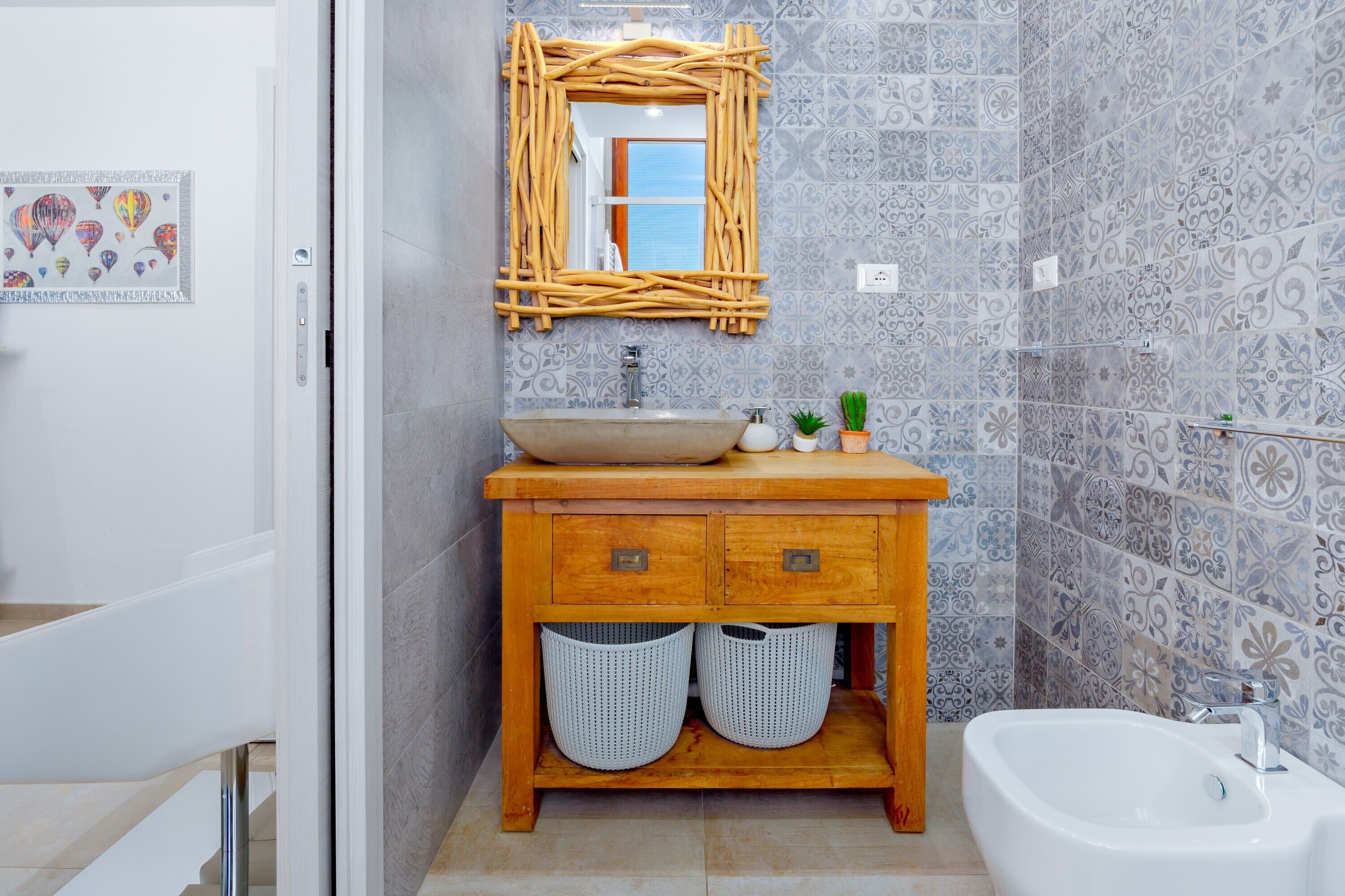 Un baño con tocador de madera, lavamanos sobre encimera, bañera independiente, azulejos estampados y espejo con marco de mimbre.