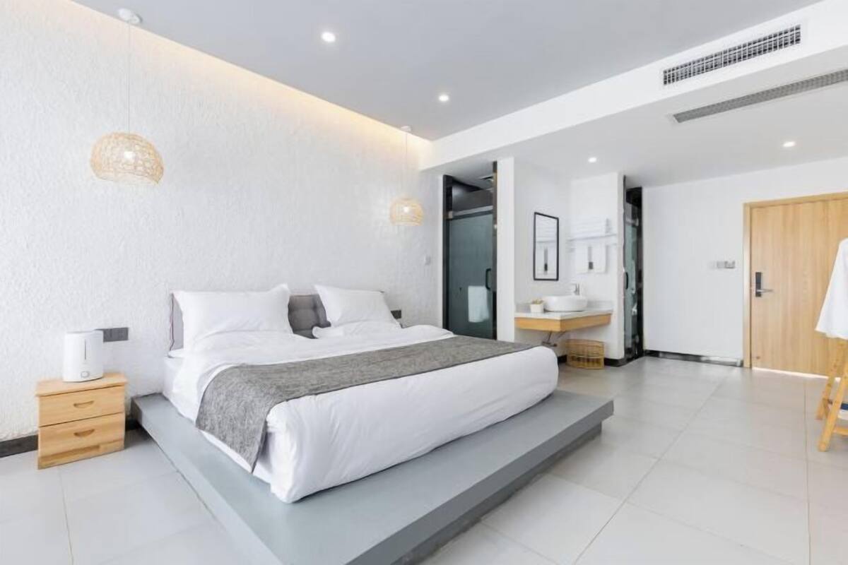 A modern hotel room with a large bed, bedside tables, a bathroom with a sink and mirror, and a wooden door.