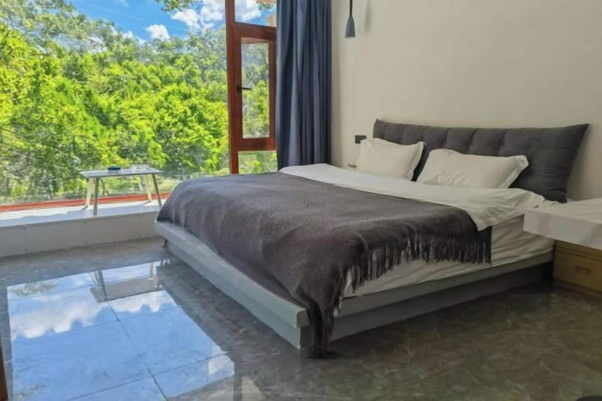 A bedroom with a bed, a nightstand, a window with a view of greenery, and a small table.