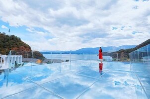 Un piso de vidrio transparente con una persona de pie, con vistas a un paisaje montañoso y una masa de agua.