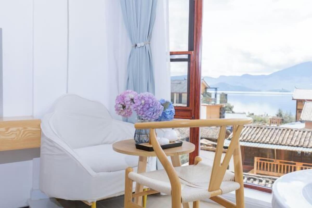 Una habitación con una silla blanca, una mesita redonda con un florero y vistas a un lago y montañas.