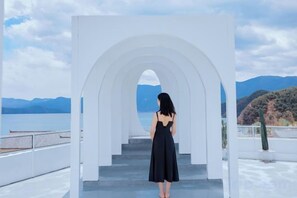 Una mujer de pie en la cima de una estructura blanca en arco, con vista a un cuerpo de agua y montañas.