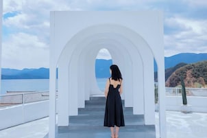 Una mujer de pie en la cima de una estructura blanca en arco, con vista a un cuerpo de agua y montañas.