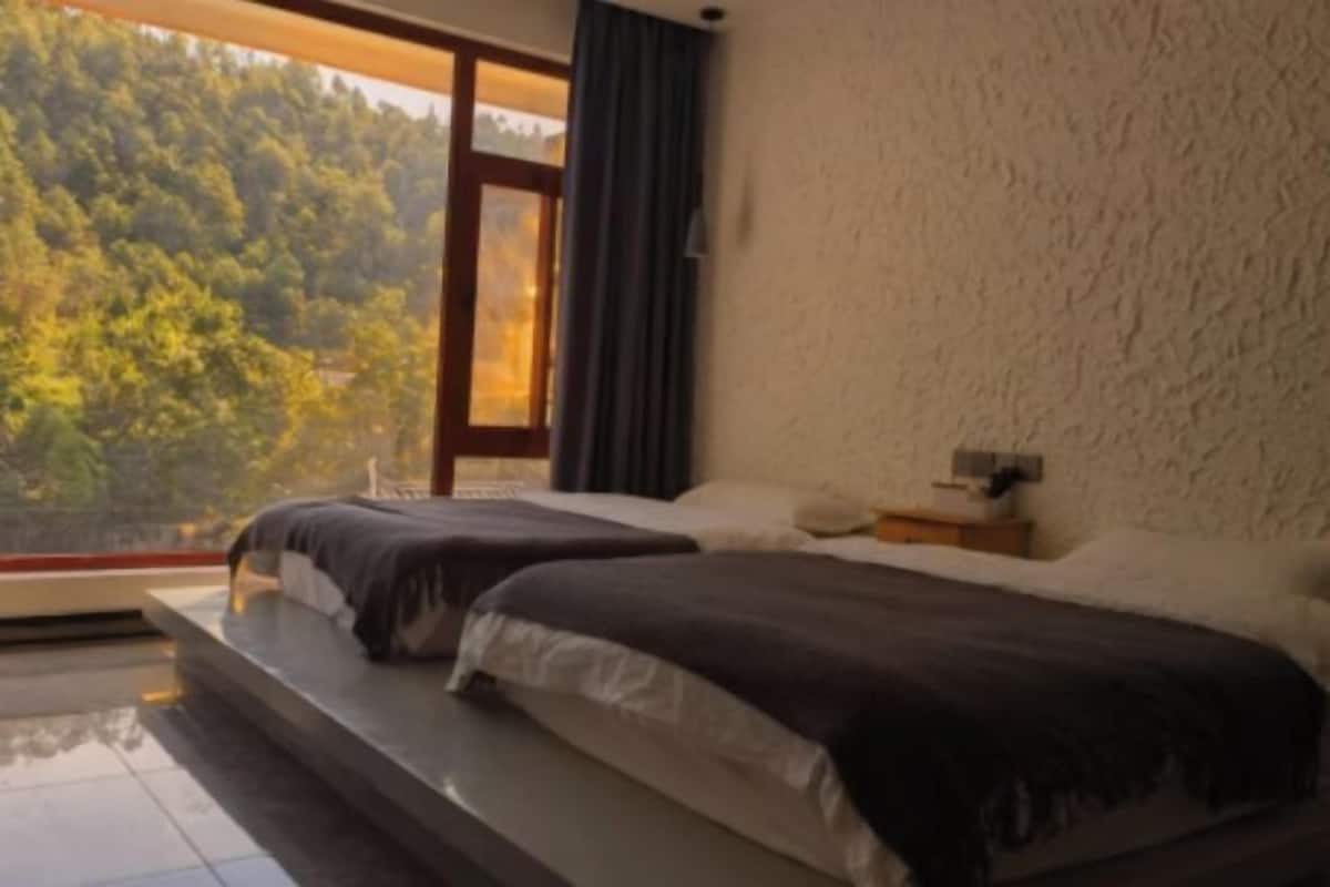 A bedroom with two beds, a large window, and a view of a forest.