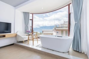 Un baño moderno con una bañera independiente, un tocador de madera y un amplio ventanal con vistas a edificios tradicionales y montañas.