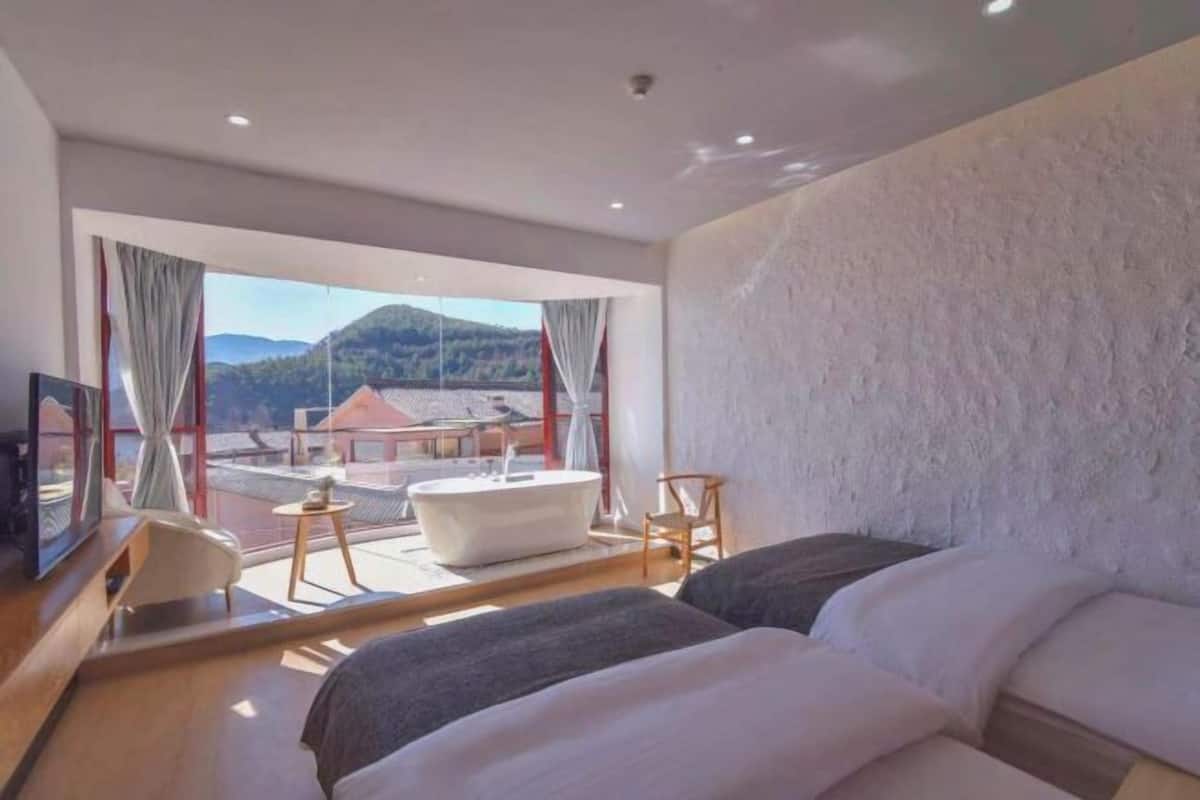 A modern hotel room with a large bed, a small desk, a bathtub, and a balcony with a view of the mountains.