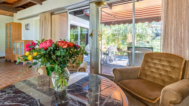 A room with a glass table, a brown sofa, a vase with flowers, and a view of the outdoors.