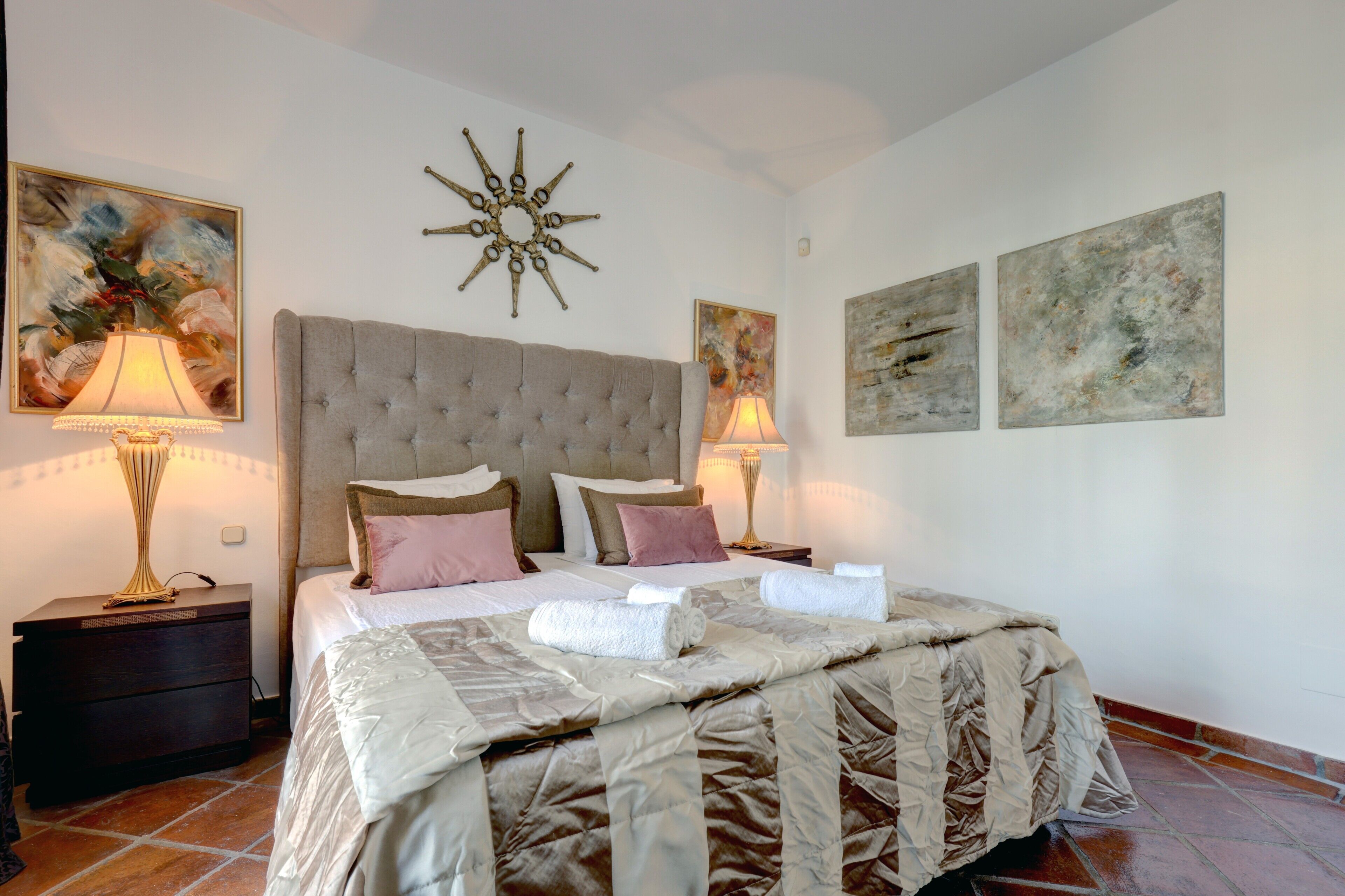 A bedroom with a tufted headboard, a large bed with multiple pillows, two bedside lamps, and artwork on the walls.
