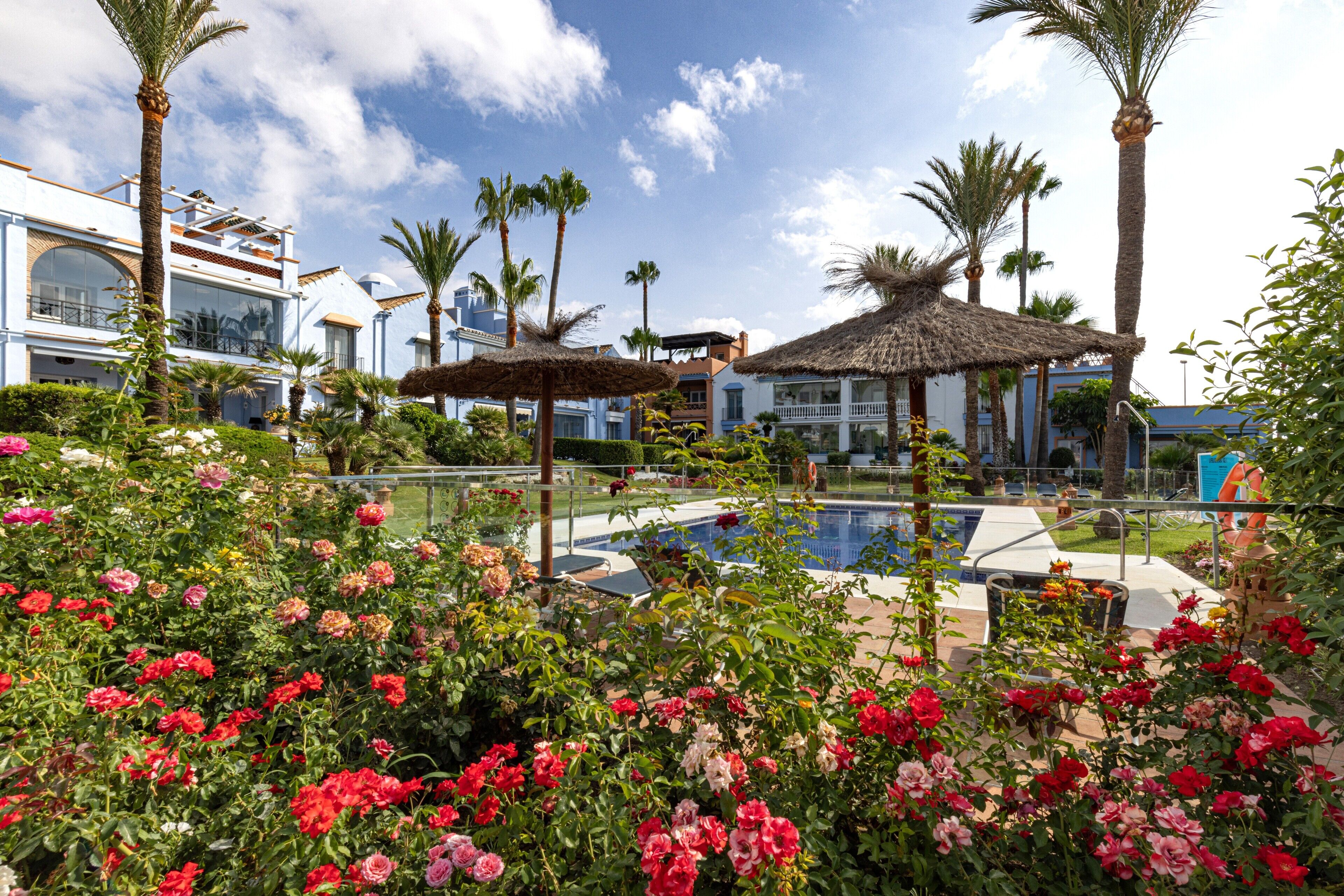 A pool area with a thatched umbrella, palm trees, and a building with a balcony.