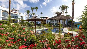 A pool area with a thatched umbrella, palm trees, and a building with a balcony.