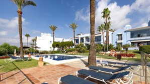 A poolside area with lounge chairs, palm trees, a swimming pool, and a building with a white facade and blue accents. - Apartment "La Perla de Marakech 5" with Sea View, Shared Pool & Wi-Fi (Casares)