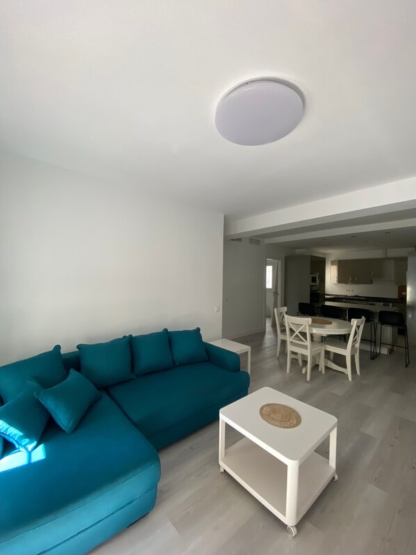 A modern living room with a teal sectional sofa, a white coffee table, and a kitchen area in the background.