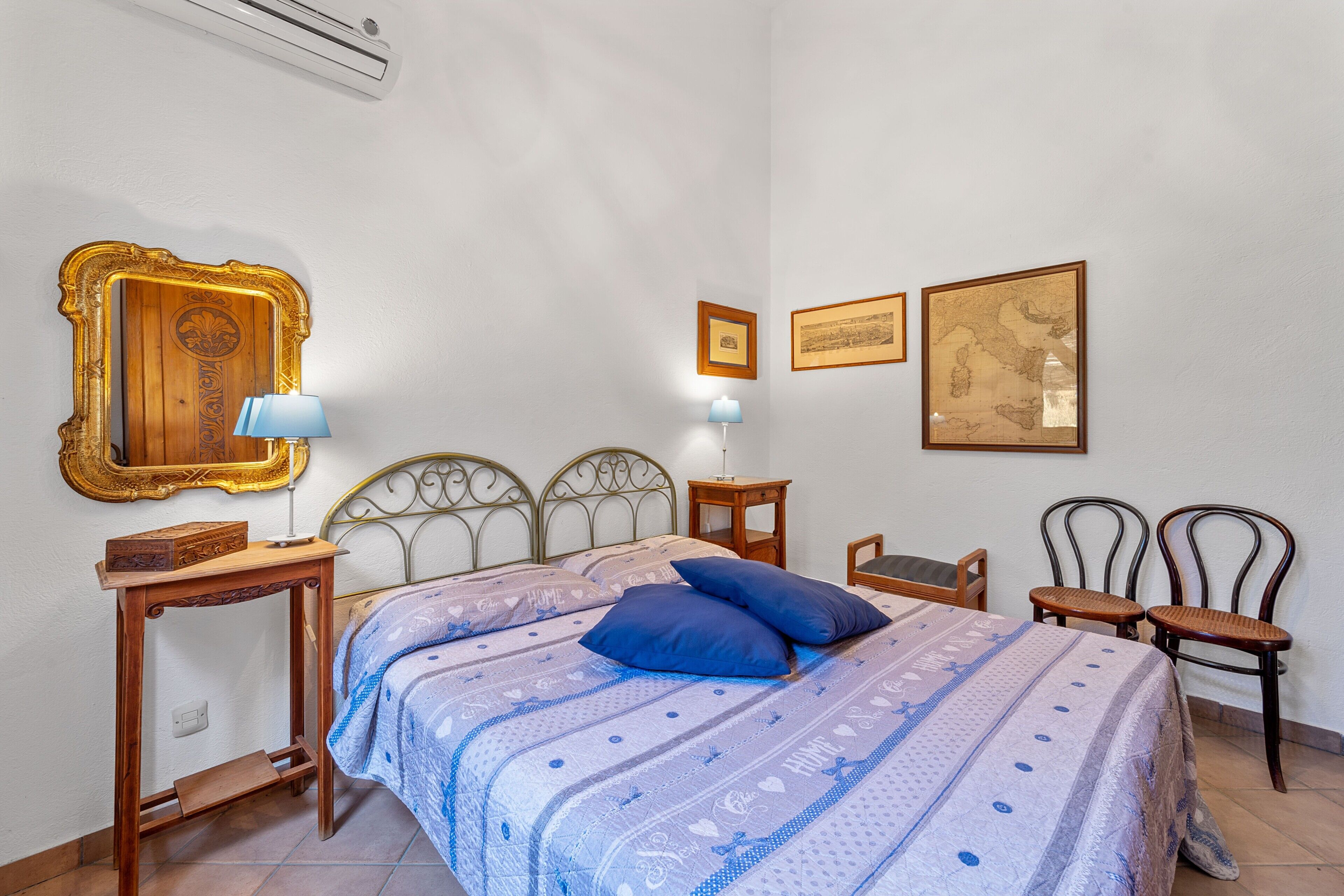 A bedroom with a bed, two bedside tables, a mirror, two wall-mounted lamps, two chairs, and framed pictures on the wall.