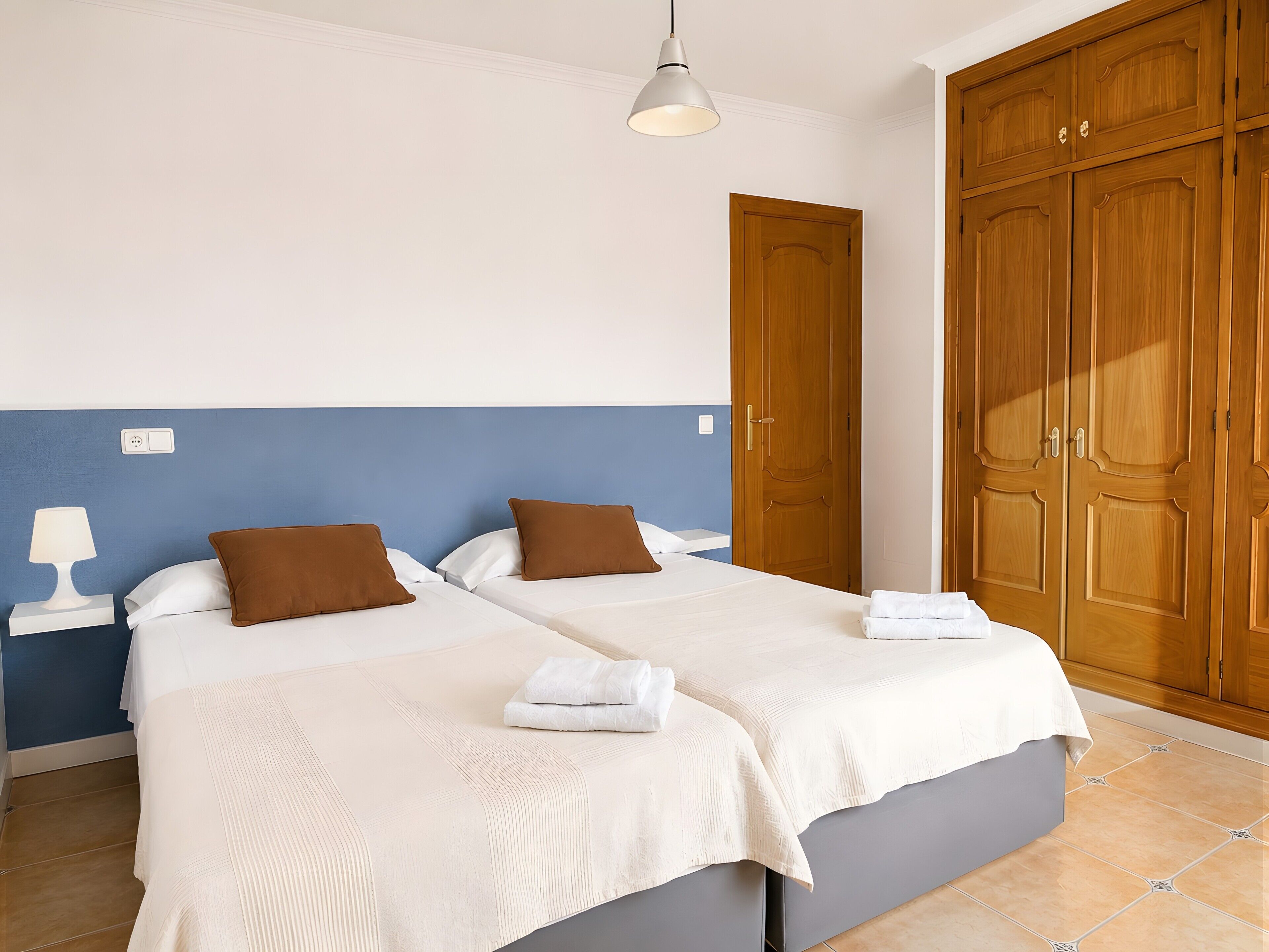 Una cama doble con ropa de cama blanca y marrón, una mesita de noche con lámpara y un armario de madera.