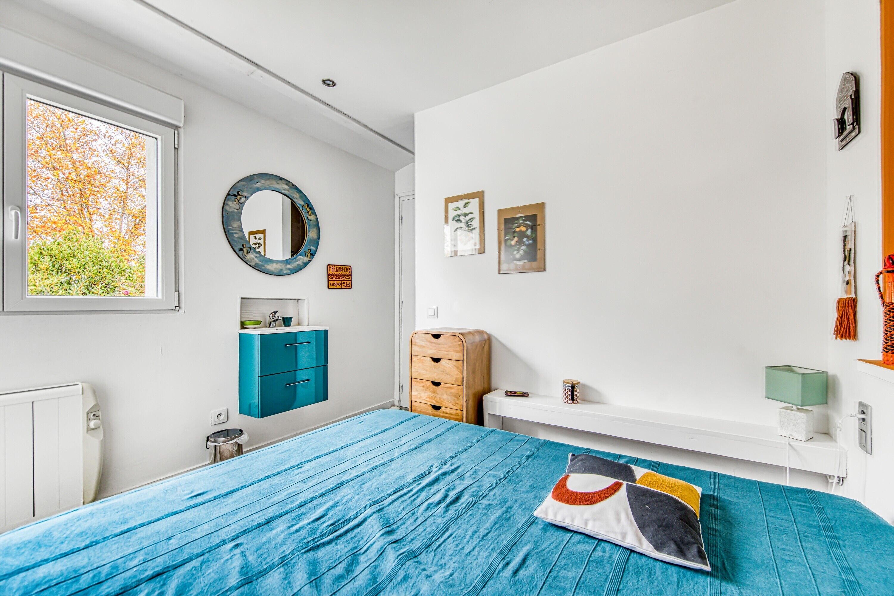 Une chambre à coucher avec un lit, une commode, un miroir et une fenêtre donnant sur des arbres.