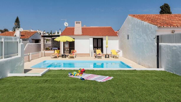 Un jardin avec une piscine, une pelouse verdoyante et un bâtiment blanc surmonté d’un toit recouvert de tuiles rouges.