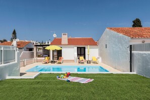 A backyard with a swimming pool, a grassy lawn, and a white building with a red-tiled roof.