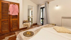 Une chambre à coucher avec un lit, une armoire en bois, une salle de bain visible à travers une porte ouverte et une fenêtre avec des rideaux.