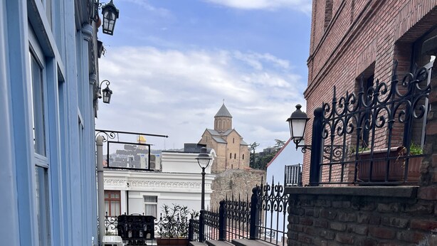 A narrow outdoor corridor with tables and chairs, a brick wall, and a building with a sign in the background. - TbilHome hostel (Tbilisi)