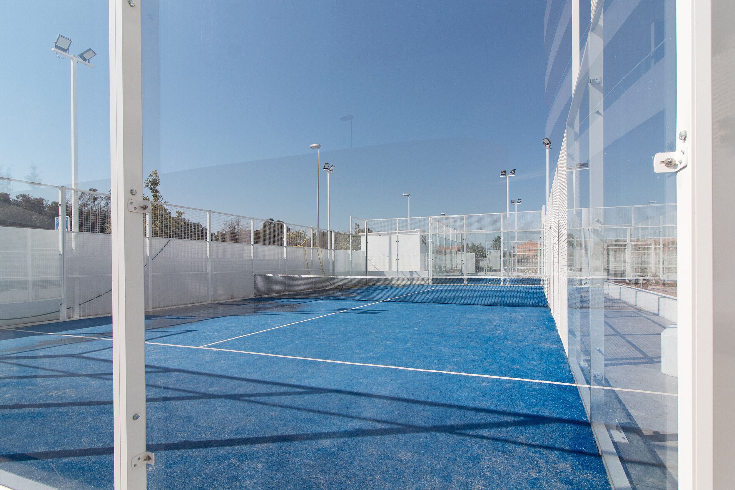 An indoor tennis court with a blue surface, enclosed by white walls and netting, and equipped with overhead lighting.