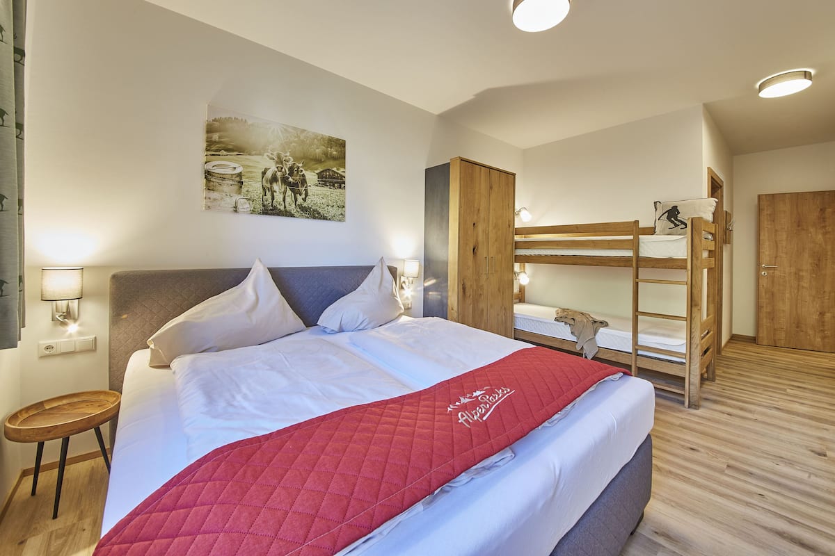 A bedroom with a bed, a wooden bunk bed, a small table, a wall-mounted lamp, and a painting.